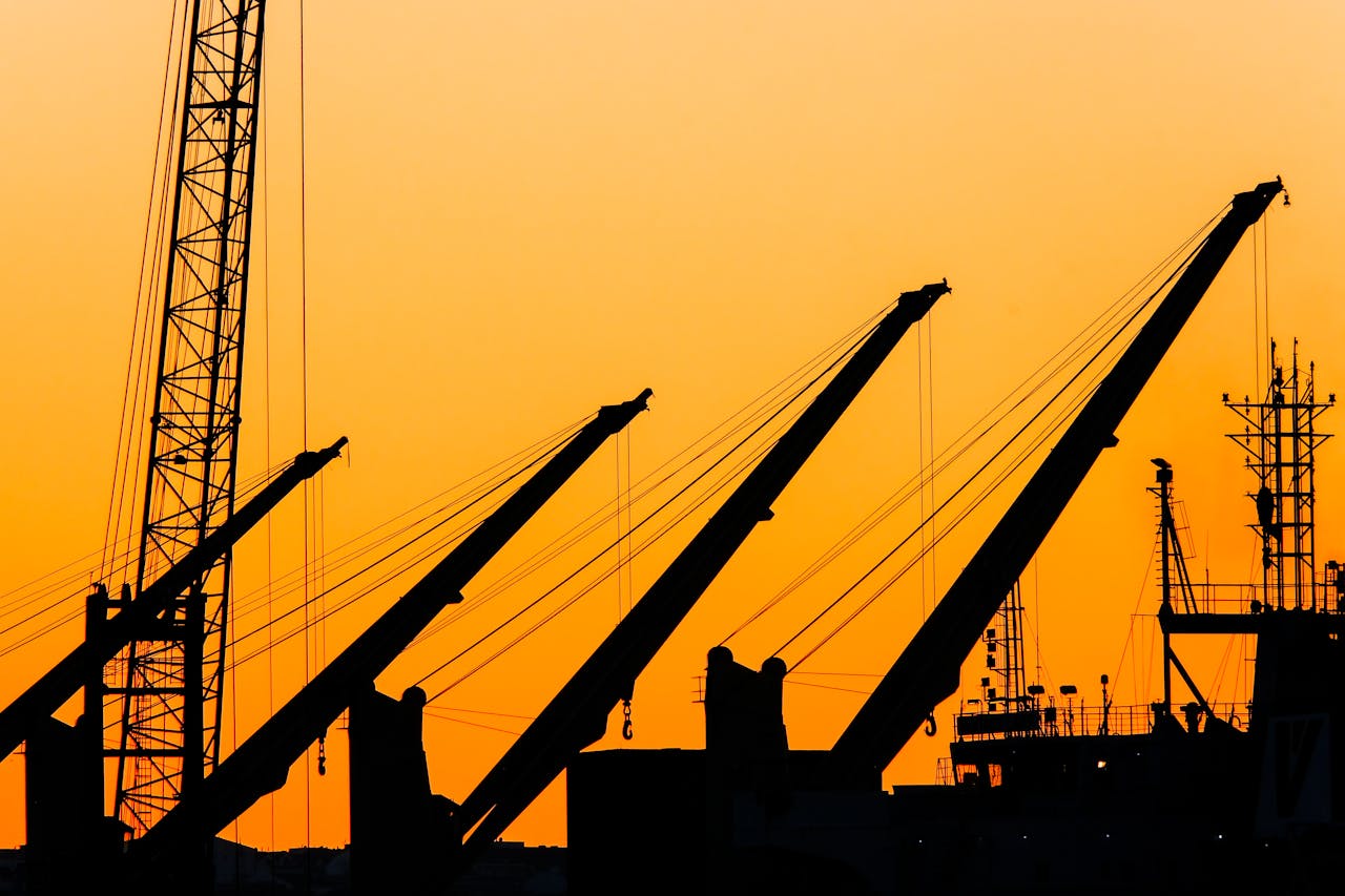 contact-img Silhouetted cranes and masts create a striking industrial skyline against a vibrant sunset backdrop.